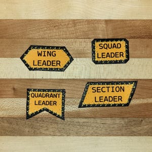 May include: Four yellow fabric patches with black borders and black text. The patches read WING LEADER, SQUAD LEADER, QUADRANT LEADER, and SECTION LEADER. Each patch features a distinct geometric shape, including a hexagon, rectangle, and pennant.