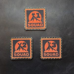 May include: Three square patches with rounded corners, featuring a black background and an orange graphic of a volcano with the numbers 1, 2, and 3 respectively. Each patch has the word SQUAD in orange and is bordered by orange stitching.