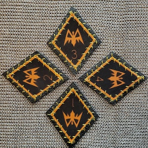May include: Four black diamond-shaped patches with orange stitching and a stylized orange emblem. Each patch has a different number: 1, 2, 3, and 4. The patches are on a textured, gray surface.