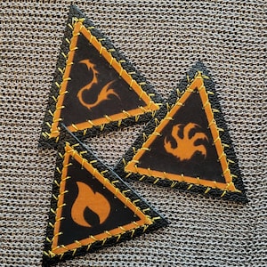 May include: Three black leather triangle patches with orange stitching and orange symbols. Each patch has a different symbol: a dragon, a flame, and a claw. The patches are on a textured, metallic surface.