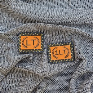 May include: Two rectangular patches with black borders and orange centers, featuring yellow stitched edges. One patch displays the letters LT, and the other displays 1LT, both surrounded by a black laurel wreath graphic printed in the center.