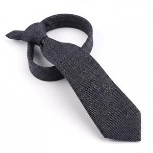 Puede incluir: Una corbata de color gris carb&oacute;n oscuro con una textura jaspeada fina y sutil. La corbata est&aacute; atada con un nudo sencillo, con la pala ancha extendi&eacute;ndose hacia abajo y el extremo estrecho doblado detr&aacute;s del nudo, mostrando un tejido uniforme.