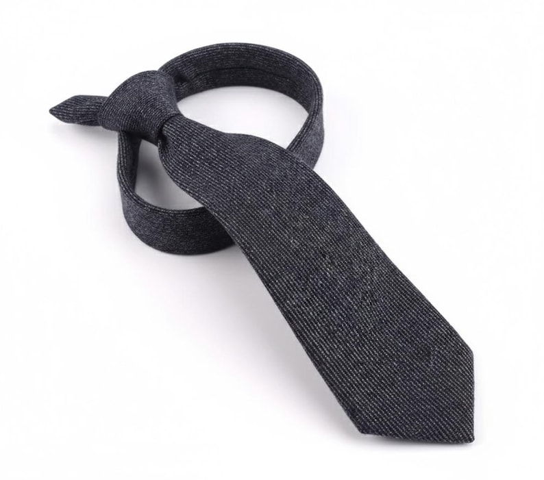 Puede incluir: Una corbata de color gris carb&oacute;n oscuro con una textura jaspeada fina y sutil. La corbata est&aacute; atada con un nudo sencillo, con la pala ancha extendi&eacute;ndose hacia abajo y el extremo estrecho doblado detr&aacute;s del nudo, mostrando un tejido uniforme.