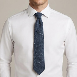 Puede incluir: Una camisa blanca de manga larga con cuello abotonado combinada con una corbata azul marino oscuro. La corbata presenta una tela texturizada con un sutil patr&oacute;n moteado que consiste en diminutos puntos blancos y gris claro sobre la superficie.