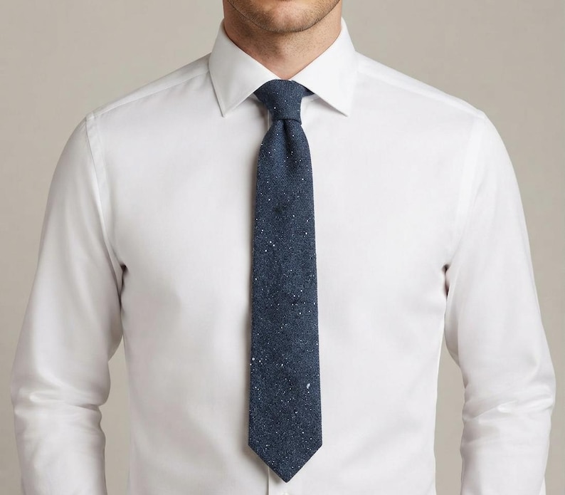 Puede incluir: Una camisa blanca de manga larga con cuello abotonado combinada con una corbata azul marino oscuro. La corbata presenta una tela texturizada con un sutil patr&oacute;n moteado que consiste en diminutos puntos blancos y gris claro sobre la superficie.