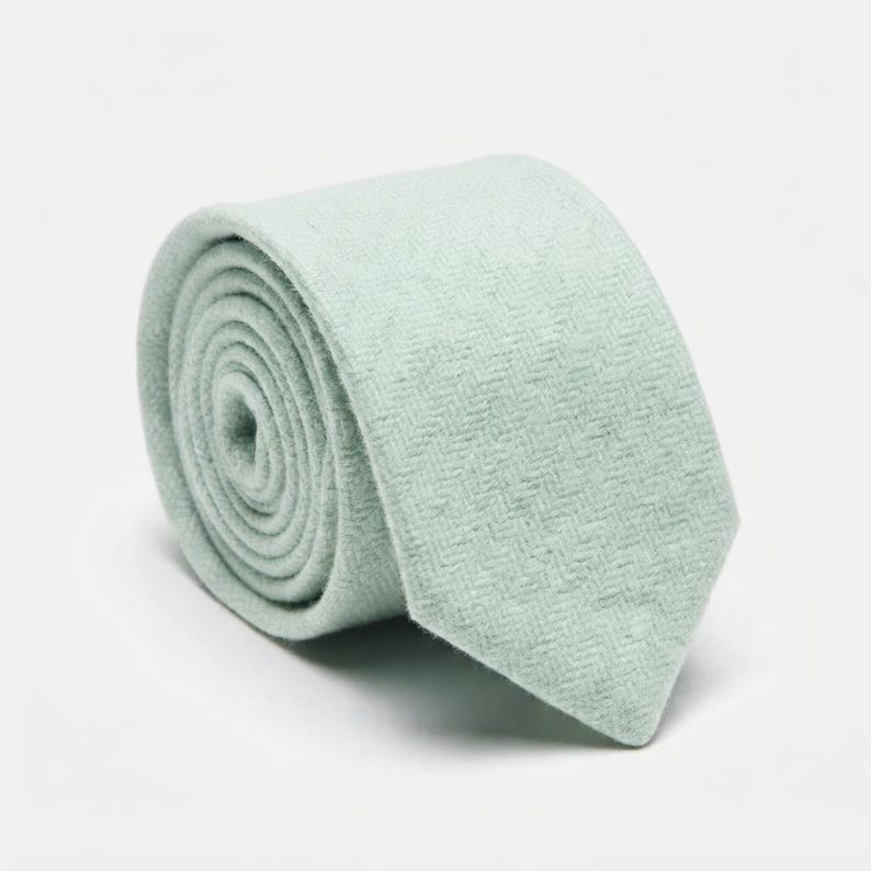 Puede incluir: Una corbata enrollada de color verde menta suave con un sutil patr&oacute;n de tejido en espiga. La tela tiene una apariencia texturizada con un acabado mate, y la corbata est&aacute; enrollada en forma circular, mostrando la pala frontal y las capas internas.