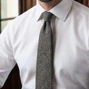 Puede incluir: Una camisa blanca de cuello abotonado combinada con una corbata gris oscuro texturizada. La corbata presenta un sutil patr&oacute;n moteado con diminutos puntos rojos y azules tejidos en la tela, creando una apariencia compleja y tonal sobre la camisa.