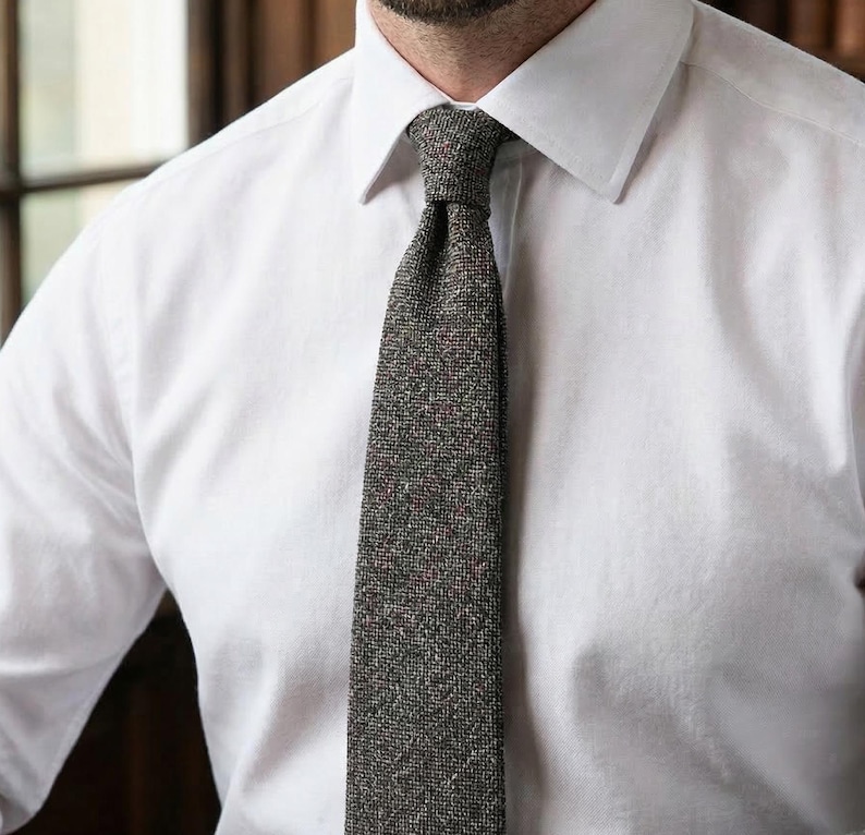 Puede incluir: Una camisa blanca de cuello abotonado combinada con una corbata gris oscuro texturizada. La corbata presenta un sutil patr&oacute;n moteado con diminutos puntos rojos y azules tejidos en la tela, creando una apariencia compleja y tonal sobre la camisa.