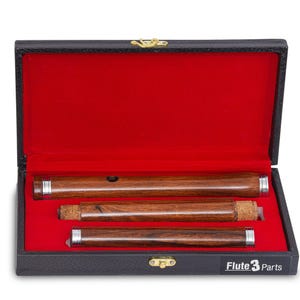 Puede incluir: Una flauta de madera en un estuche negro con interior de terciopelo rojo. La flauta está en tres partes, con detalles plateados. El estuche está etiquetado como "Flute 3 Parts".