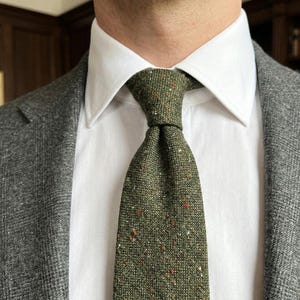 Puede incluir: Una corbata de color verde oliva con un patr&oacute;n texturizado y moteado que presenta peque&ntilde;as motas rojas, blancas y marr&oacute;n oscuro. La corbata se lleva con una camisa blanca de cuello r&iacute;gido y un blazer gris carb&oacute;n texturizado, creando un estilo formal.