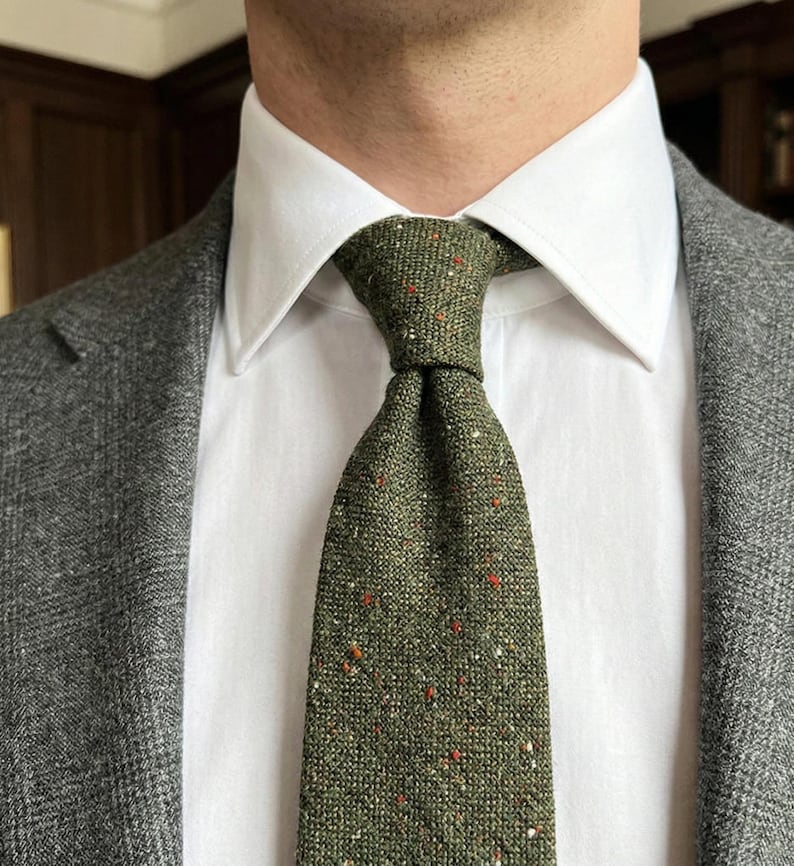 Puede incluir: Una corbata de color verde oliva con un patr&oacute;n texturizado y moteado que presenta peque&ntilde;as motas rojas, blancas y marr&oacute;n oscuro. La corbata se lleva con una camisa blanca de cuello r&iacute;gido y un blazer gris carb&oacute;n texturizado, creando un estilo formal.