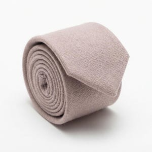 Puede incluir: Una corbata enrollada de color malva apagado con un sutil patr&oacute;n de espiga. La tela tiene una textura suave similar al fieltro y la corbata est&aacute; enrollada en forma circular con el extremo puntiagudo descansando sobre la parte superior.