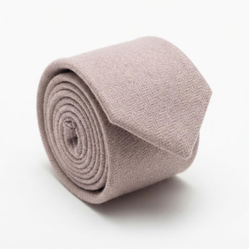 Puede incluir: Una corbata enrollada de color malva apagado con un sutil patr&oacute;n de espiga. La tela tiene una textura suave similar al fieltro y la corbata est&aacute; enrollada en forma circular con el extremo puntiagudo descansando sobre la parte superior.