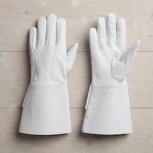 Puede incluir: Un par de guantes de cuero blanco estilo guantelete con puños largos, cinco dedos y detalles de costuras en el dorso de la mano. Los guantes tienen un acabado liso y un diseño estructurado que se extiende más allá de la muñeca.