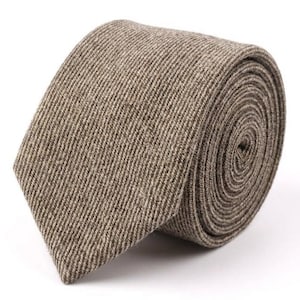 Puede incluir: Una corbata con un patr&oacute;n de espiga texturizado en tonos marr&oacute;n claro y beige. La tela tiene un tejido visible con una apariencia ligeramente moteada. La corbata est&aacute; enrollada en una bobina apretada, mostrando el extremo puntiagudo.