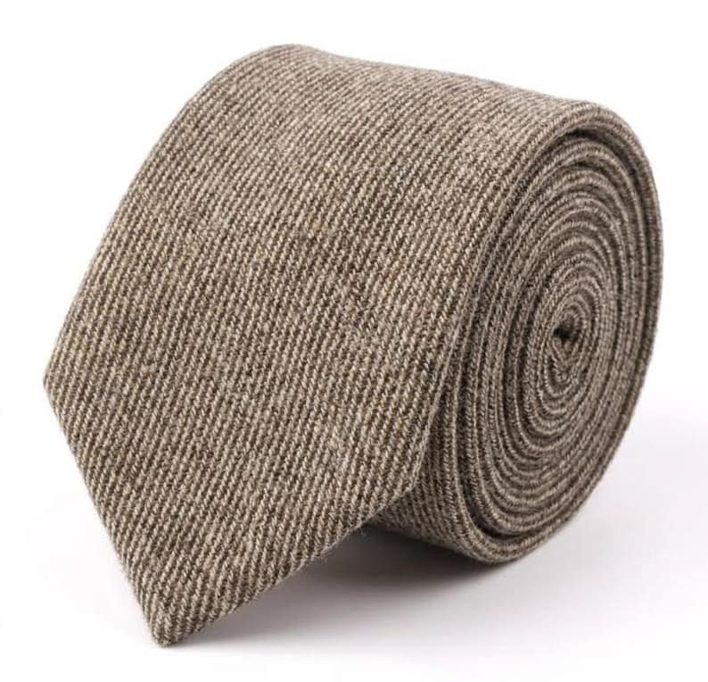 Puede incluir: Una corbata con un patr&oacute;n de espiga texturizado en tonos marr&oacute;n claro y beige. La tela tiene un tejido visible con una apariencia ligeramente moteada. La corbata est&aacute; enrollada en una bobina apretada, mostrando el extremo puntiagudo.