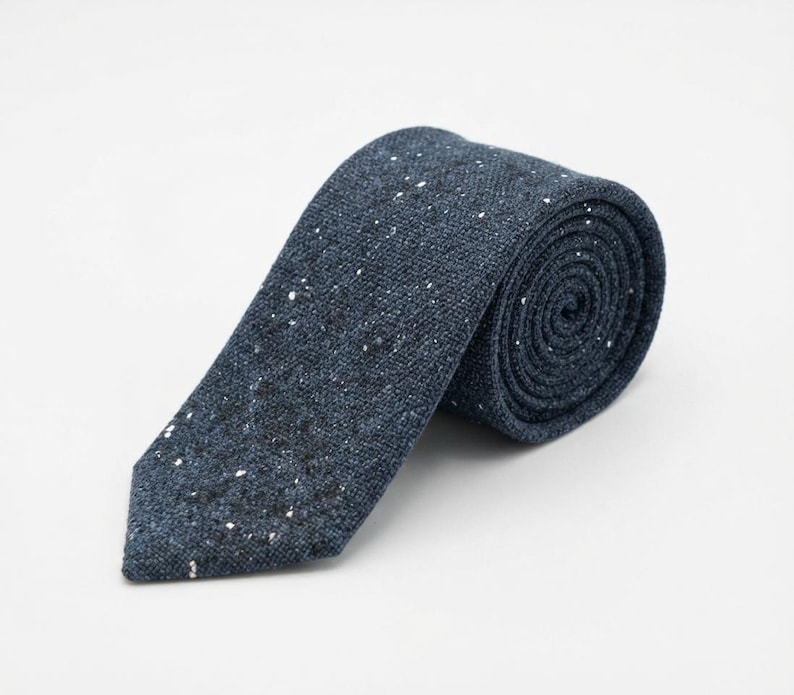 Puede incluir: Una corbata azul oscuro con un patr&oacute;n texturizado y moteado con peque&ntilde;as motas blancas y negras por toda la tela. La corbata est&aacute; enrollada en forma de espiral, mostrando claramente el material tejido y el extremo puntiagudo de la corbata.