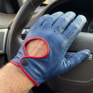 May include: Blue driving gloves with red trim and a snap closure. The gloves have small perforations for breathability. The gloves are on a steering wheel.