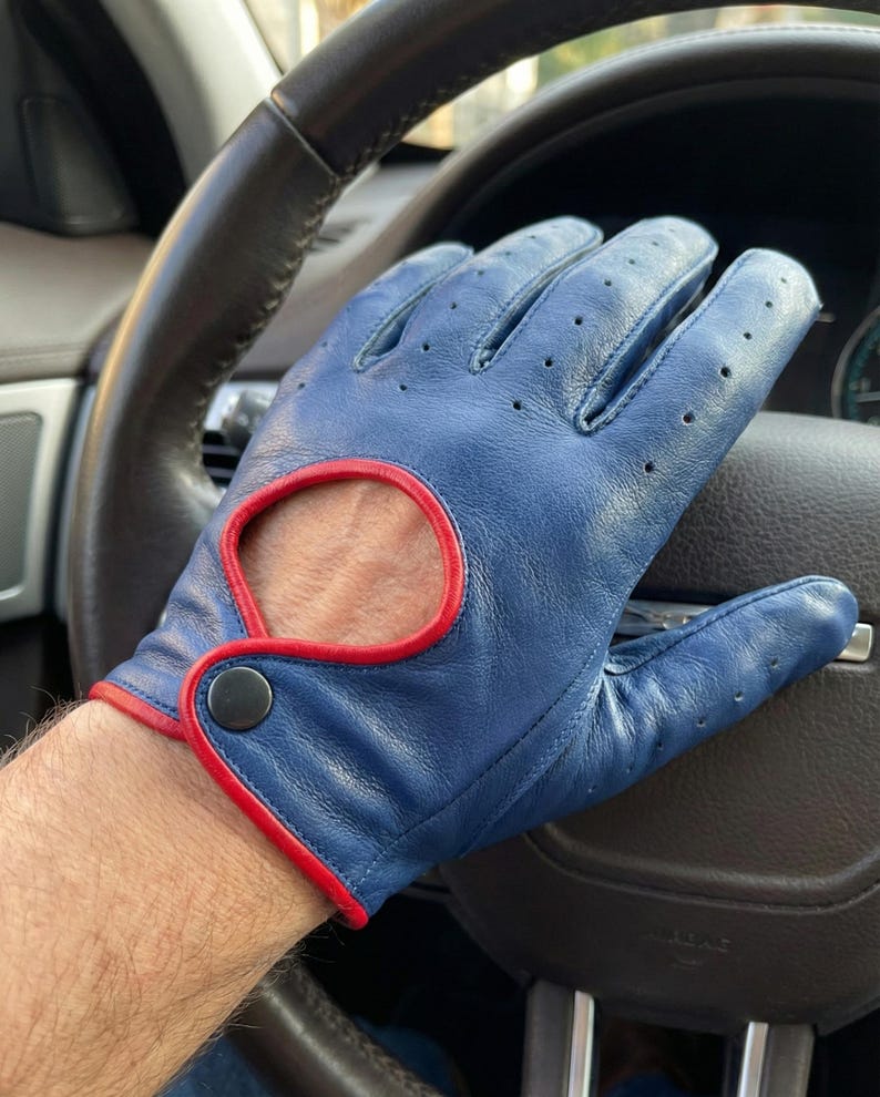 May include: Blue driving gloves with red trim and a snap closure. The gloves have small perforations for breathability. The gloves are on a steering wheel.