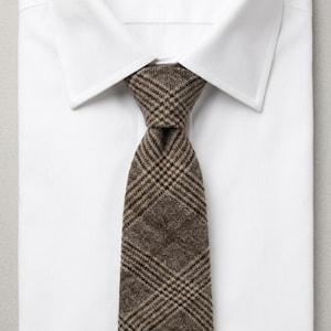 Puede incluir: Una corbata de cuadros marr&oacute;n y beige con una apariencia tejida y texturizada, anudada y colocada sobre una camisa de vestir blanca lisa con cuello. La corbata presenta un patr&oacute;n de cuadros diagonales cl&aacute;sico en tonos tierra, contrastando con la tela.