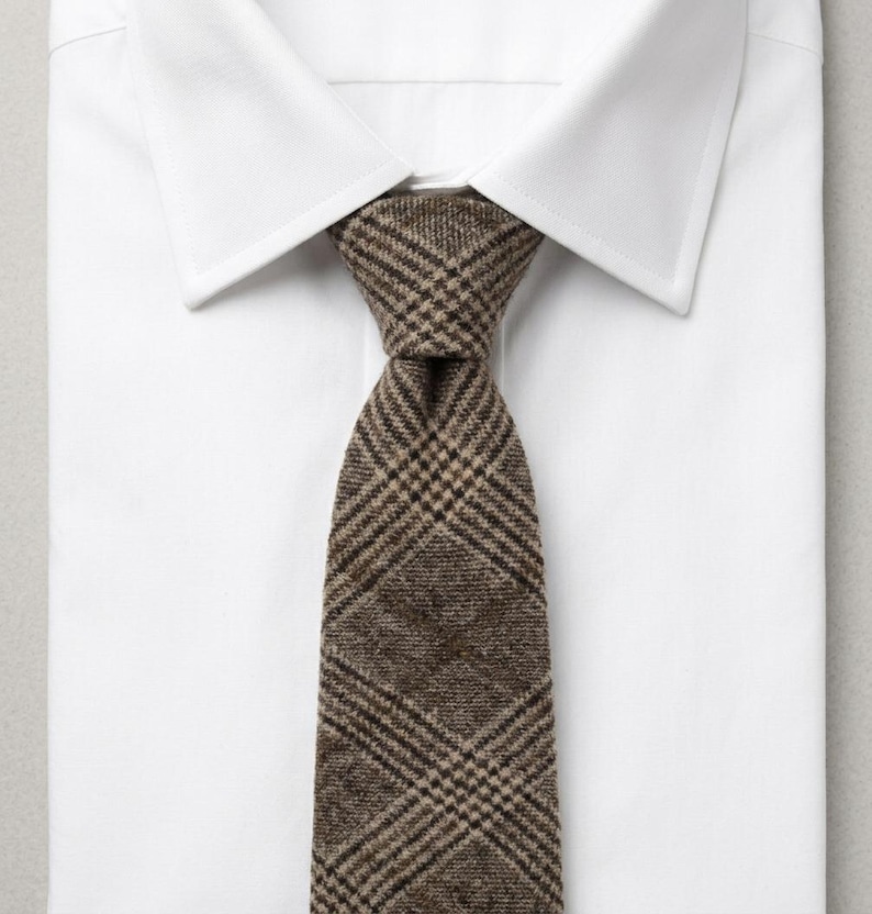 Puede incluir: Una corbata de cuadros marr&oacute;n y beige con una apariencia tejida y texturizada, anudada y colocada sobre una camisa de vestir blanca lisa con cuello. La corbata presenta un patr&oacute;n de cuadros diagonales cl&aacute;sico en tonos tierra, contrastando con la tela.