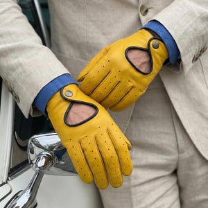 May include: Bright yellow driving gloves with black trim and small perforations. The gloves have a distinctive cut-out design and are secured with a button closure. The gloves are worn with a light tan blazer and a blue shirt.