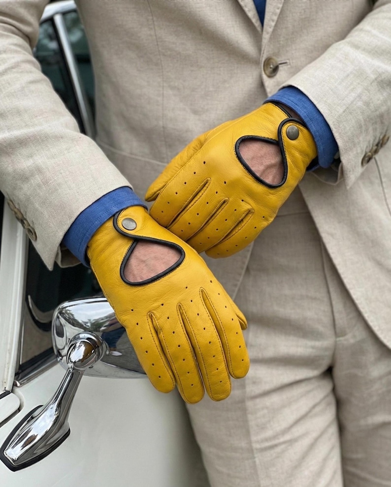 May include: Bright yellow driving gloves with black trim and small perforations. The gloves have a distinctive cut-out design and are secured with a button closure. The gloves are worn with a light tan blazer and a blue shirt.