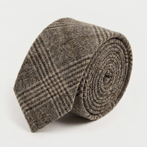 Puede incluir: Una corbata enrollada en forma circular, con una tela marr&oacute;n texturizada y un patr&oacute;n de cuadros en marr&oacute;n oscuro y beige. El material tiene un acabado tejido visible y la corbata se muestra con el extremo puntiagudo visible al frente.