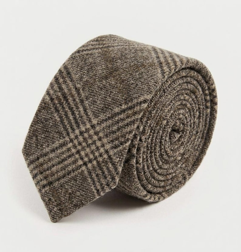 Puede incluir: Una corbata enrollada en forma circular, con una tela marr&oacute;n texturizada y un patr&oacute;n de cuadros en marr&oacute;n oscuro y beige. El material tiene un acabado tejido visible y la corbata se muestra con el extremo puntiagudo visible al frente.