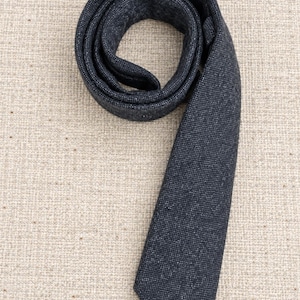 Puede incluir: Una corbata de color gris carb&oacute;n oscuro con un acabado de tela texturizada y moteada. La corbata est&aacute; enrollada en forma circular en la parte superior, con el extremo puntiagudo extendi&eacute;ndose hacia abajo, mostrando un tejido uniforme.