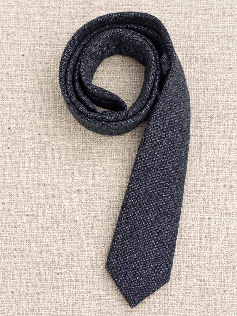 Puede incluir: Una corbata de color gris carb&oacute;n oscuro con un acabado de tela texturizada y moteada. La corbata est&aacute; enrollada en forma circular en la parte superior, con el extremo puntiagudo extendi&eacute;ndose hacia abajo, mostrando un tejido uniforme.
