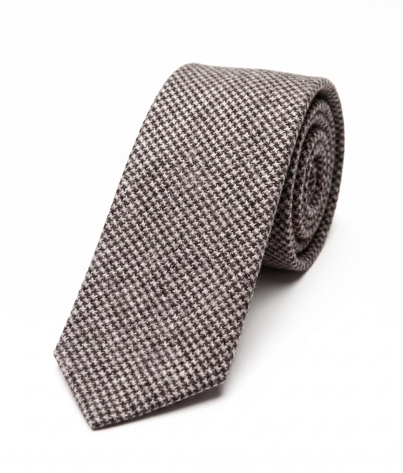 Puede incluir: Una corbata con un patr&oacute;n de pata de gallo en marr&oacute;n y blanco. La tela tiene una apariencia tejida y texturizada. La corbata est&aacute; enrollada en forma circular, mostrando claramente el extremo puntiagudo y la estructura del material enrollado.