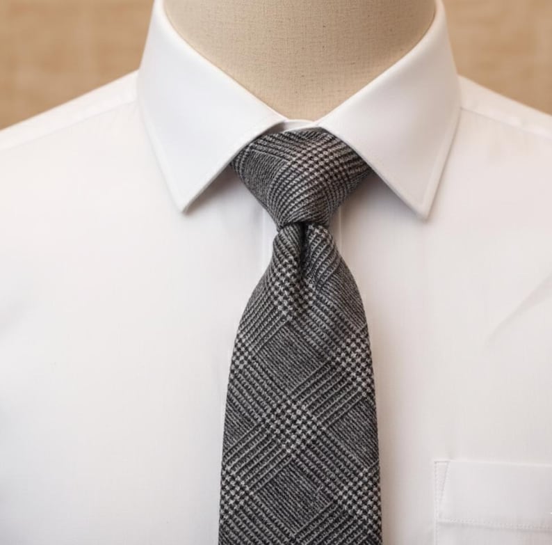 Puede incluir: Una camisa blanca de cuello r&iacute;gido con una corbata gris que presenta un detallado patr&oacute;n de cuadros en blanco y negro. La corbata est&aacute; anudada cuidadosamente en el centro, mostrando un tejido texturizado y un dise&ntilde;o geom&eacute;trico cl&aacute;sico.