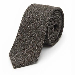 Puede incluir: Una corbata de color marr&oacute;n oscuro con un tejido texturizado y moteado que presenta peque&ntilde;as motas blancas, bronceadas y negras. La corbata est&aacute; enrollada en forma circular, mostrando su material tejido y su extremo puntiagudo.