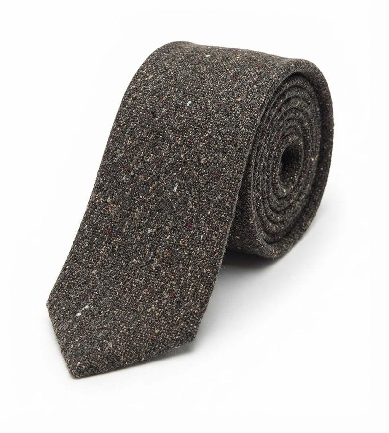 Puede incluir: Una corbata de color marr&oacute;n oscuro con un tejido texturizado y moteado que presenta peque&ntilde;as motas blancas, bronceadas y negras. La corbata est&aacute; enrollada en forma circular, mostrando su material tejido y su extremo puntiagudo.