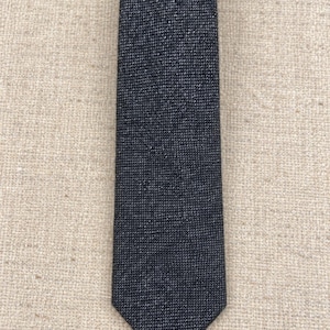 Puede incluir: Una corbata de color gris carb&oacute;n oscuro con un patr&oacute;n de tejido fino y texturizado. La corbata tiene una punta cl&aacute;sica y una construcci&oacute;n de tela densa y uniforme en toda su longitud, mostrando un aspecto jaspeado sutil en el material.
