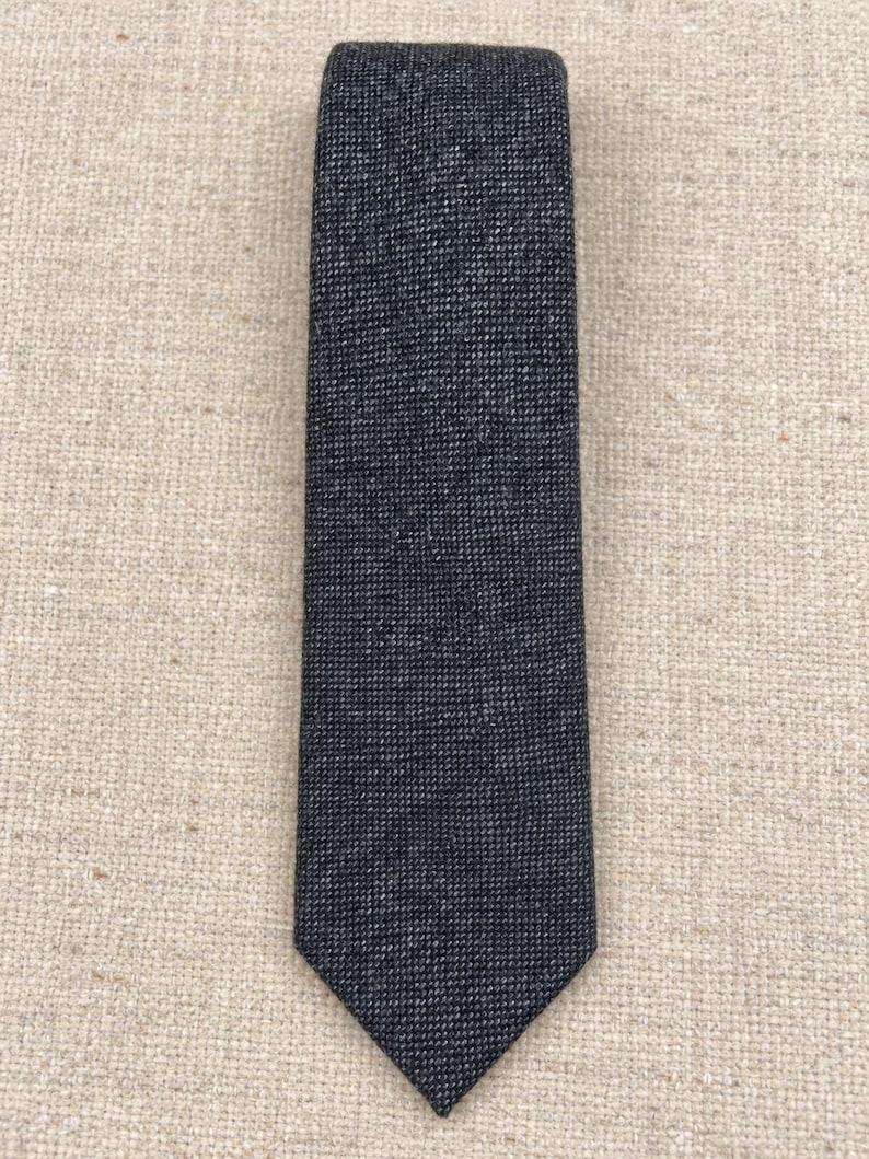 Puede incluir: Una corbata de color gris carb&oacute;n oscuro con un patr&oacute;n de tejido fino y texturizado. La corbata tiene una punta cl&aacute;sica y una construcci&oacute;n de tela densa y uniforme en toda su longitud, mostrando un aspecto jaspeado sutil en el material.