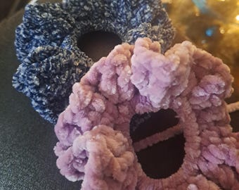 May include: Two plush scrunchies in the image. One is a deep navy blue, and the other is a soft lavender color. The scrunchies are made of a fuzzy, textured material, likely velvet or a similar fabric. These hair accessories are designed to hold hair in place.