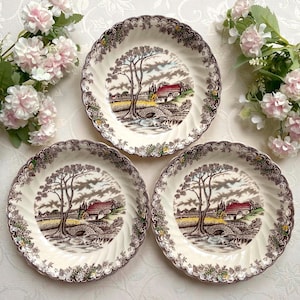 May include: Three cream-colored ceramic plates featuring a brown transferware landscape scene of a cottage, a stone bridge, and a large tree. The scalloped edges are decorated with a brown floral pattern and small yellow accents.
