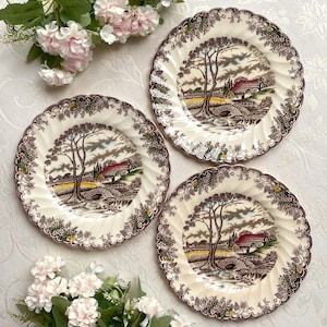 May include: Three round ceramic plates featuring a scalloped edge and a brown transferware landscape pattern. The design depicts a cottage, a stone bridge, and a large tree. The rim is decorated with a floral border in brown, green, and yellow.