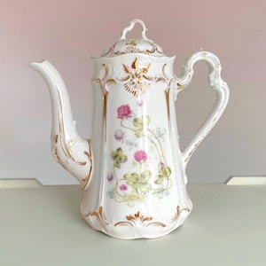 May include: A white porcelain teapot featuring a delicate floral pattern of pink clover blossoms and green leaves. The teapot has a curved handle, a long spout, and a matching lid, all accented with intricate gold trim along the edges and base.