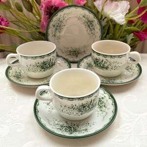 Puede incluir: Un juego de tazas de té blancas con un diseño floral verde y ribete. El juego incluye tres tazas, tres platillos y un plato más grande. Las tazas tienen asas y los platillos son redondos.