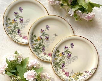 May include: Three cream-colored plates with gold rims are arranged on a white surface. Each plate features a floral design with purple, yellow, and pink flowers. The plates are surrounded by pink and white flowers.