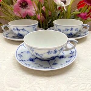 Puede incluir: Cuatro tazas y platillos blancos con diseños florales azules. Las tazas tienen asas curvas y se asientan sobre platillos a juego. El fondo presenta flores rosas y blancas.