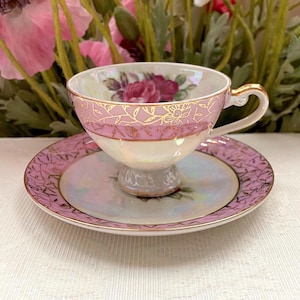 Puede incluir: Un juego de taza de té y platillo de porcelana con acabado iridiscente, una banda rosa suave y patrones de filigrana floral dorada. El interior de la taza muestra un motivo de rosa pintada, mientras que el platillo refleja el diseño rosa y dorado.