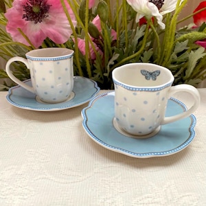 Può includere: Due tazze da tè e piattini in ceramica bianca con un motivo a pois azzurri. I piattini e i bordi delle tazze sono azzurri con un bordo smerlato. Una tazza ha un disegno a farfalla all'interno.