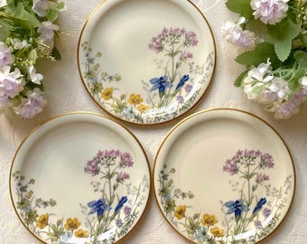 May include: Three cream-colored plates with a gold rim and floral design. The plates feature a central cluster of purple flowers, blue irises, and yellow buttercups. The floral pattern is repeated on each plate, creating a cohesive set.