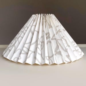 May include: A conical lampshade featuring a white base with a repeating light gray abstract floral pattern. The lampshade has a structured, accordion-style pleated design that radiates from the smaller top opening to the wider bottom edge.