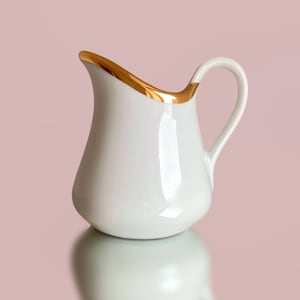Puede incluir: Una jarra de leche de cerámica blanca con un acabado liso y brillante y un asa curva. El borde superior de la jarra presenta un adorno de color dorado metálico que sigue el contorno del pico vertedor y se extiende hacia el asa.