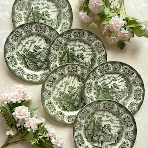 May include: A collection of seven green and white decorative plates. Each plate displays a detailed landscape scene with buildings and trees. The plates have a scalloped edge with a floral pattern. Pink flowers are arranged around the plates.